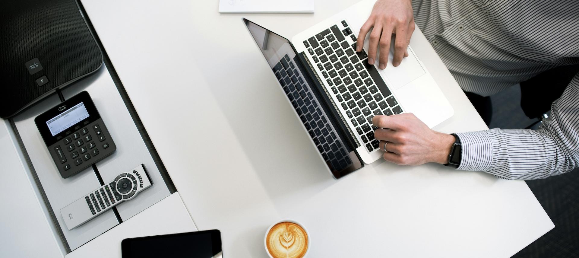 Man at laptop with coffee, pen and notebook wearing business shirt.
